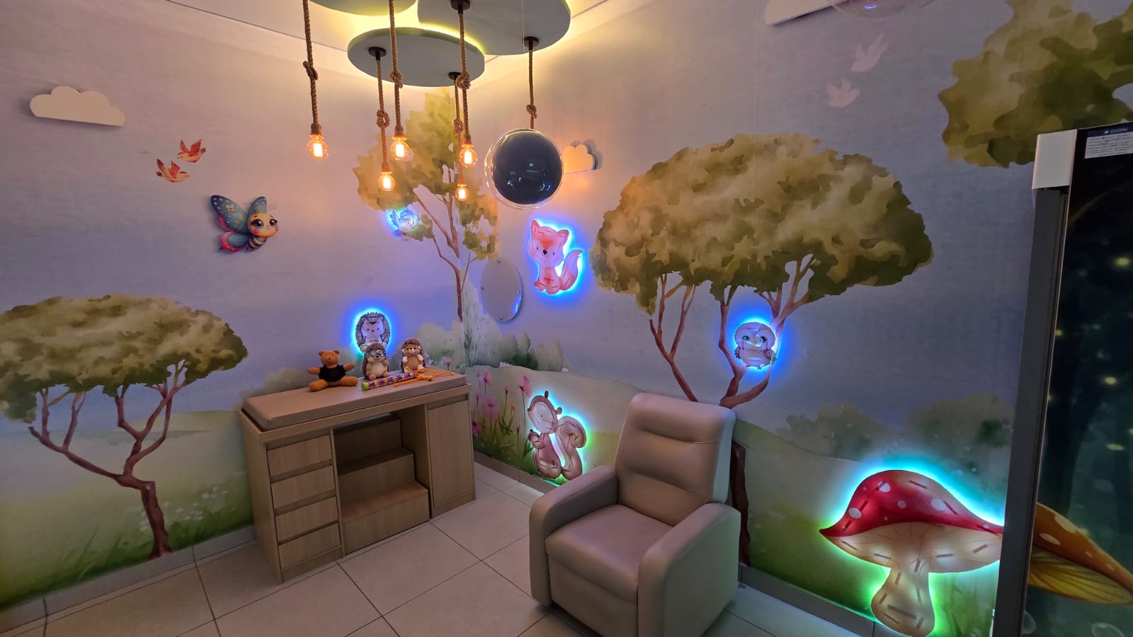 Sala de aplicação da Acolle com mural de floresta encantada, animais iluminados, poltrona e luminárias suspensas