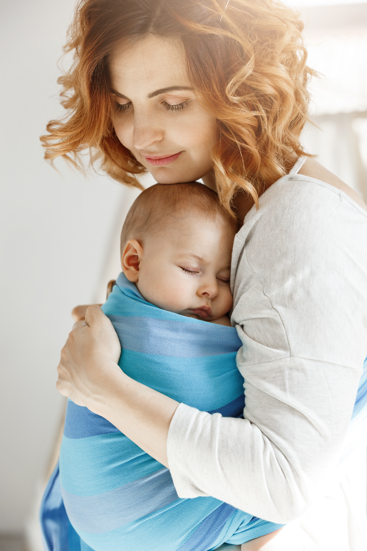 Mãe ruiva abraçando bebê adormecido em sling azul, transmitindo acolhimento e ternura
